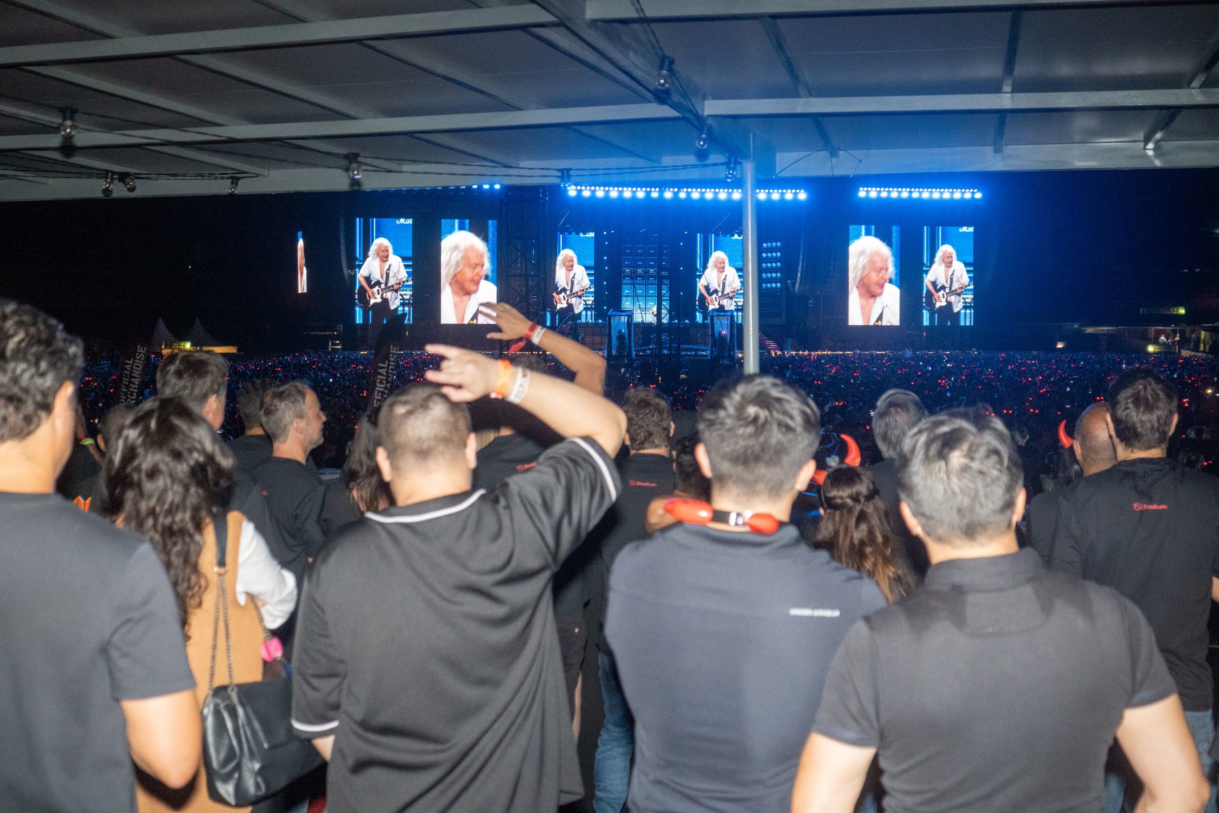 Interior de camarote coberto em estádio, com grupo de convidados de costas acompanhando o show e vista aberta para o palco e para o público na pista.