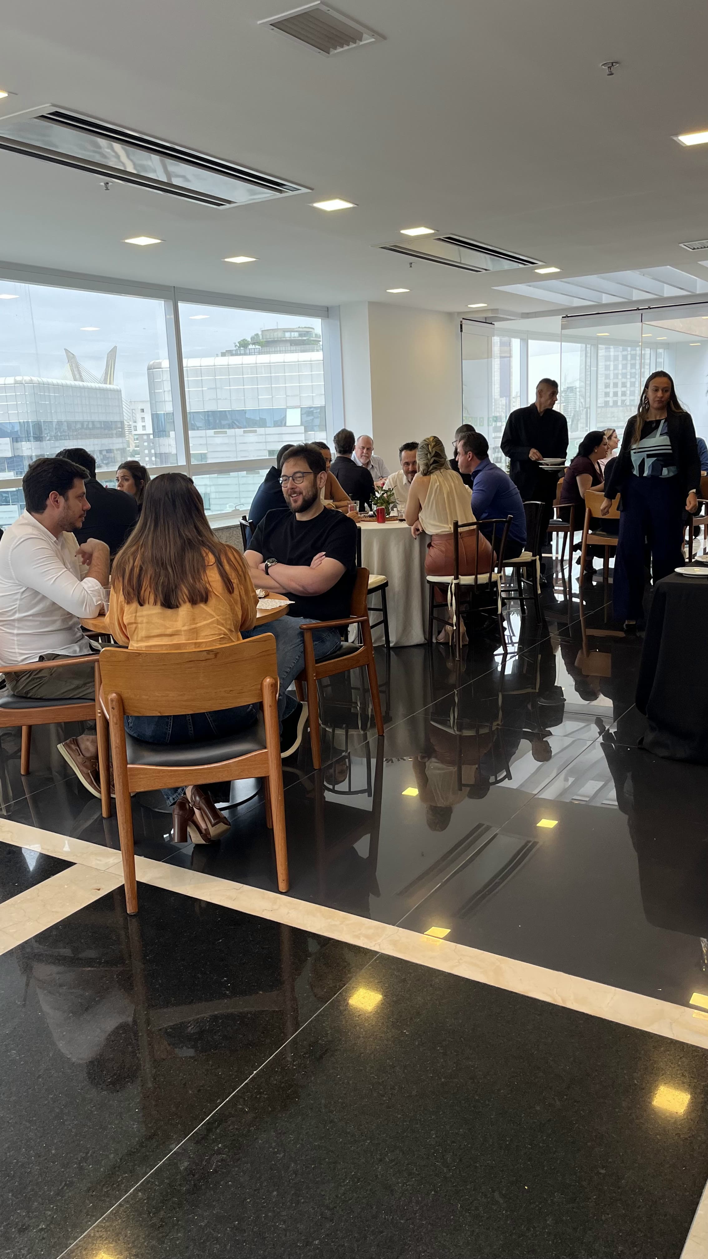Convidados sentados em mesas redondas dentro de espaço corporativo envidraçado com vista para a cidade, com equipe de serviço circulando.