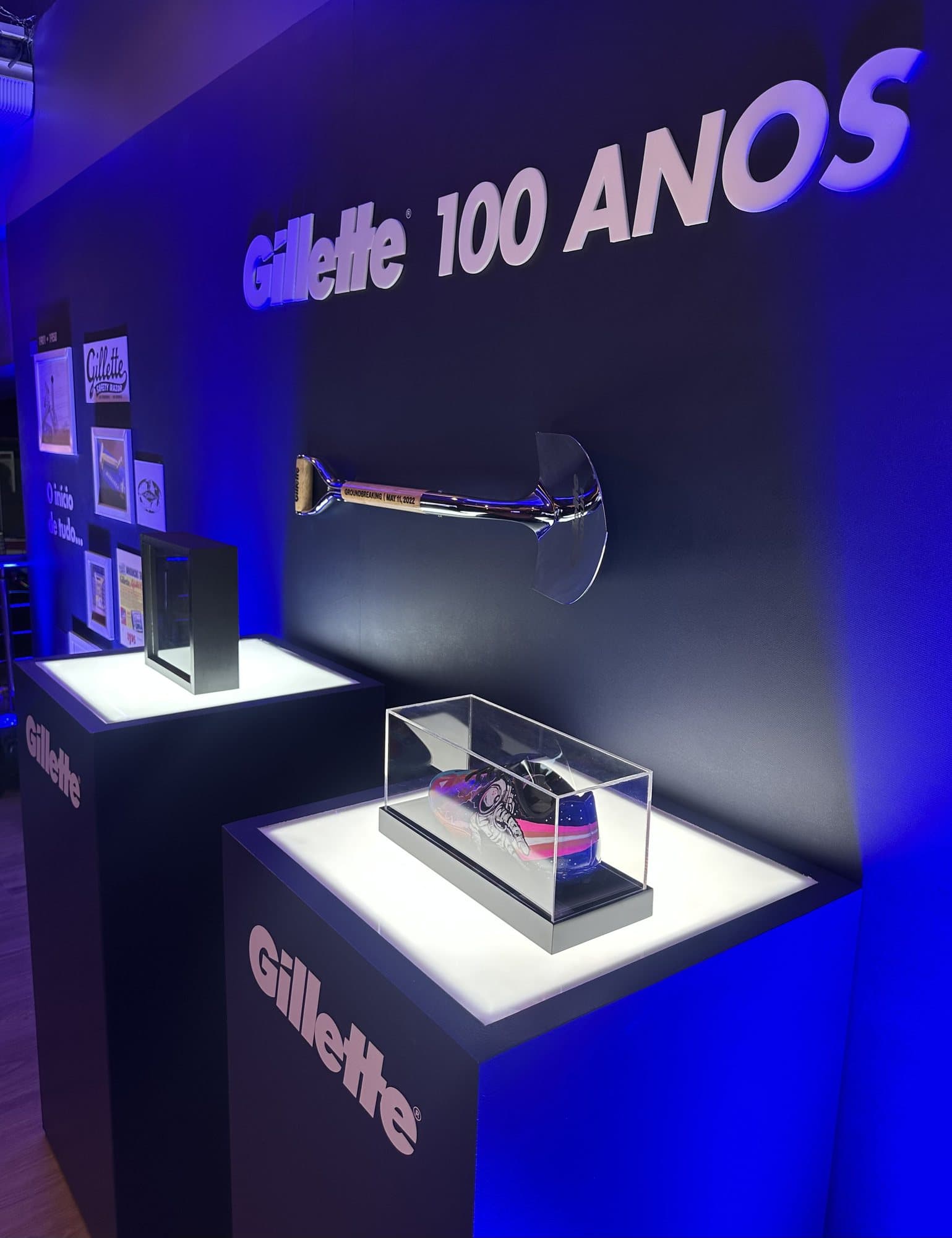 Parede da exposição de 100 anos da Gillette com o letreiro da marca iluminado em azul, uma peça cromada em destaque e expositores com objetos em caixas de acrílico.
