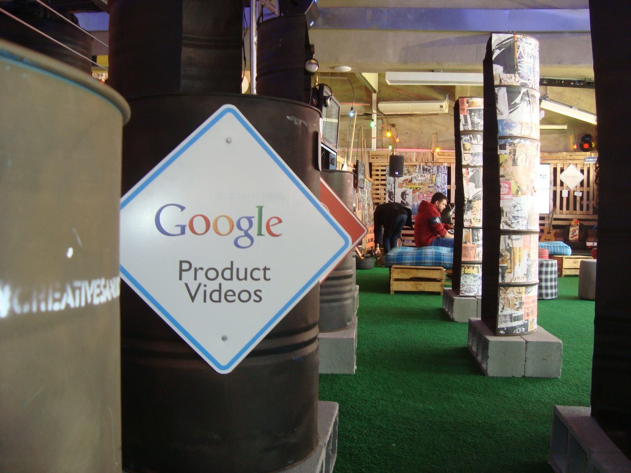 Lounge construído com paredes de pallets de madeira, videowall exibindo uma interface do Google, grama sintética, pufes xadrez e mobiliário de estar.
