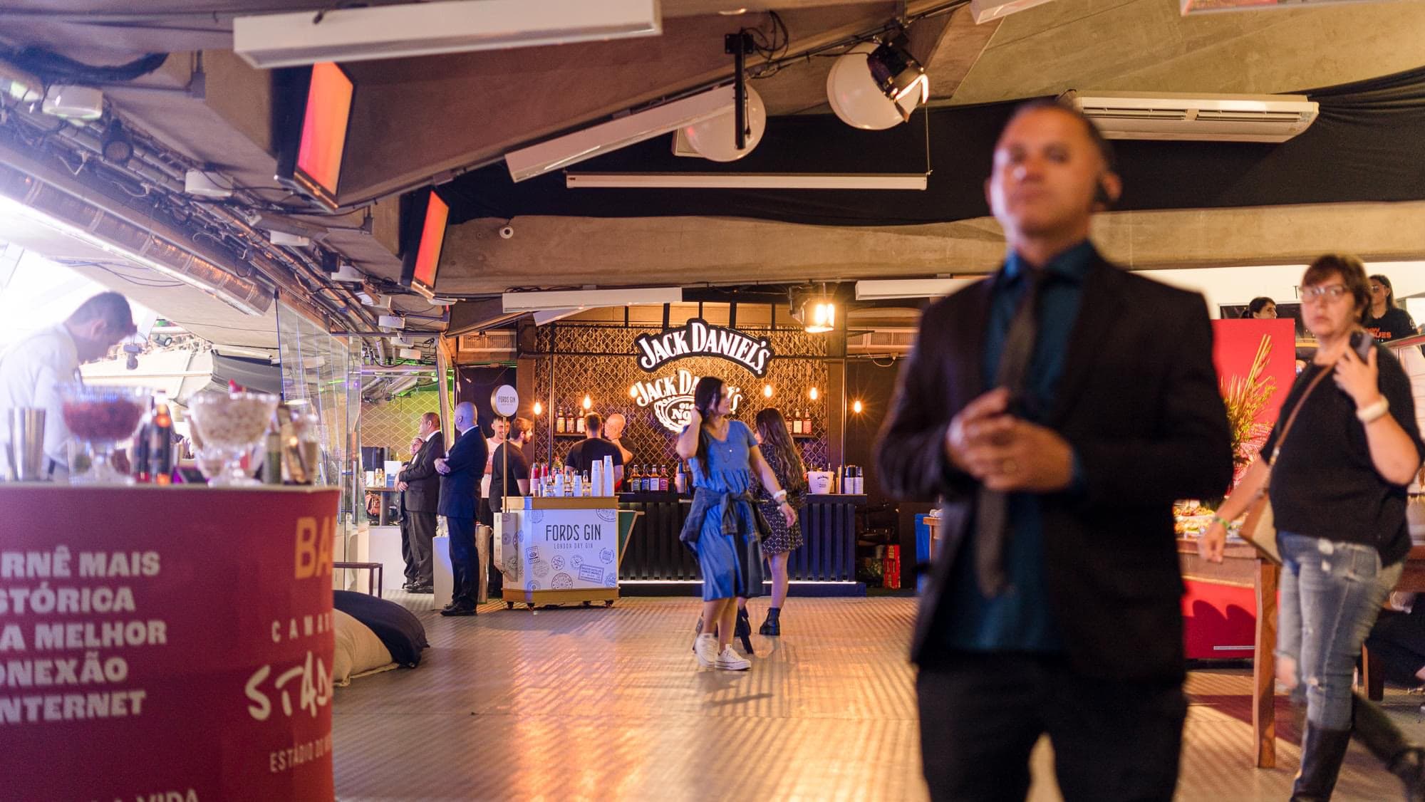 Bar cenografado com a marca Jack Daniel's em painel iluminado ao fundo, carrinho de gim em primeiro plano e convidados circulando pelo camarote.