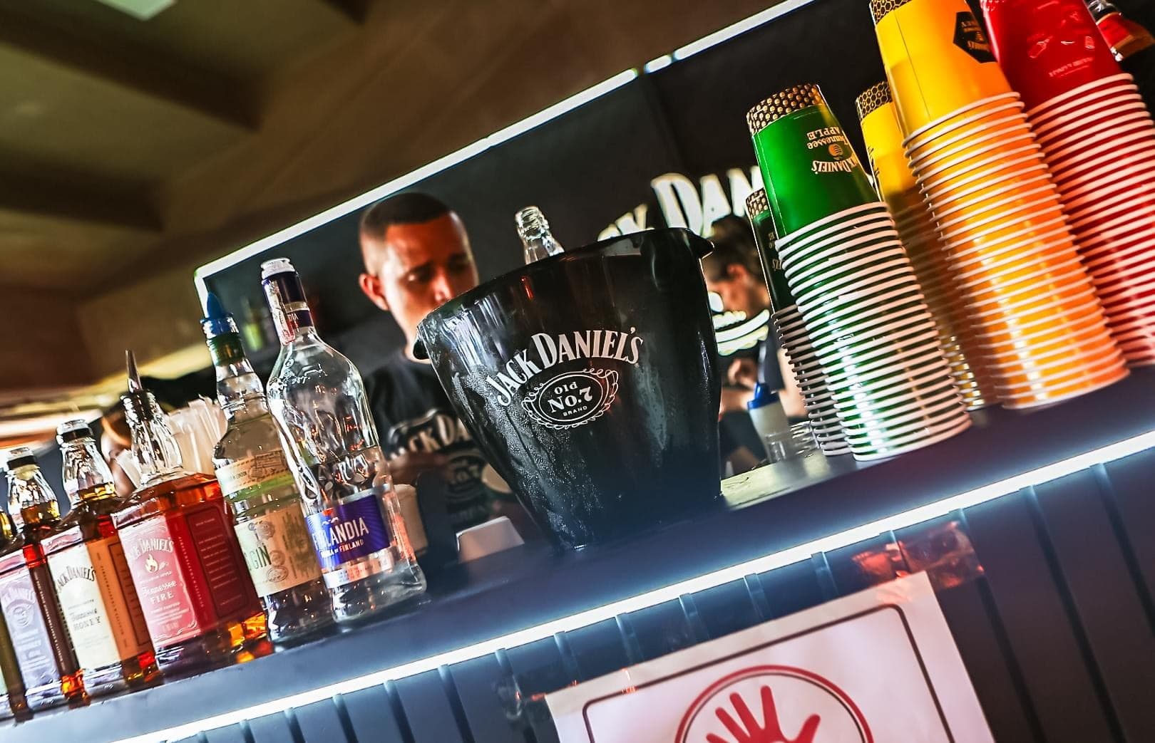 Balcão de bar da ativação de marca no camarote, com garrafas, balde de gelo personalizado e copos coloridos empilhados, e bartender preparando drinks ao fundo.
