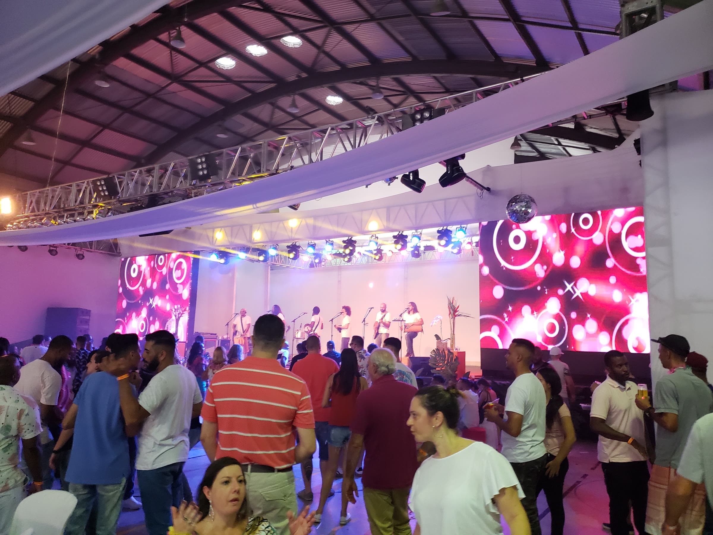 Área coberta com estrutura de treliça, refletores móveis e dois painéis de LED, com banda tocando no palco e público reunido na pista.